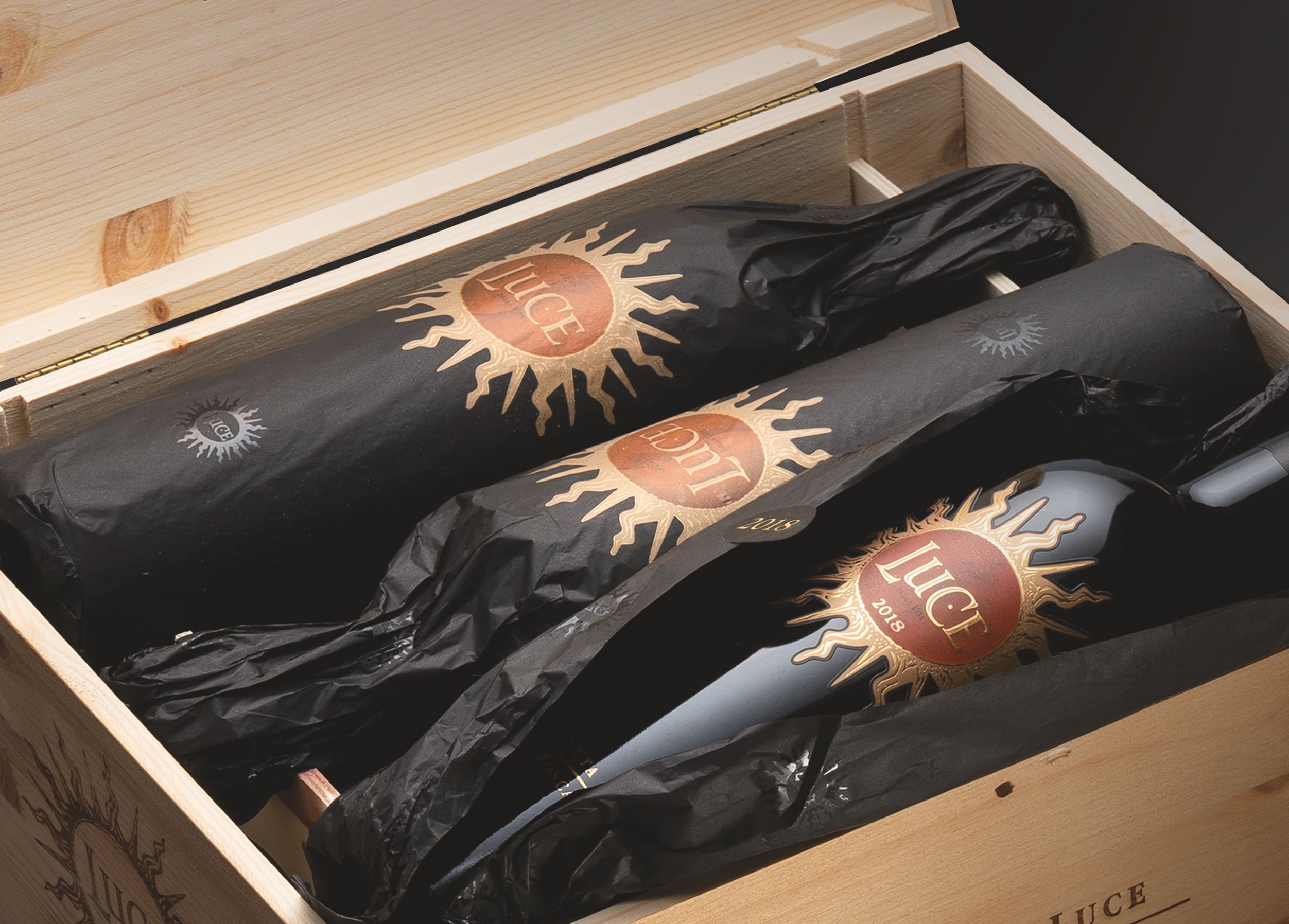 Original Holzkiste Luce Drei Flaschen Wein sind in schwarzes Papier gewickelt und in einer Holzbox angeordnet. Die Flaschen tragen Etiketten mit einer Sonnensymbolik und Jahreszahlen.