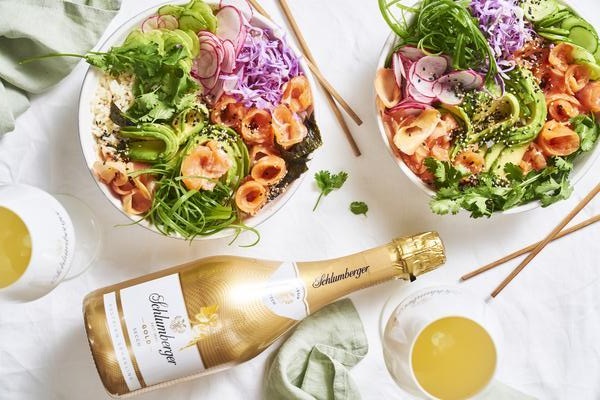 Zwei bunte Schalen mit Salat, Fisch und Gemüse sind nebeneinander angerichtet. Eine Flasche Schaumwein sowie Gläser mit Getränken und Essstäbchen liegen ebenfalls auf dem Tisch.