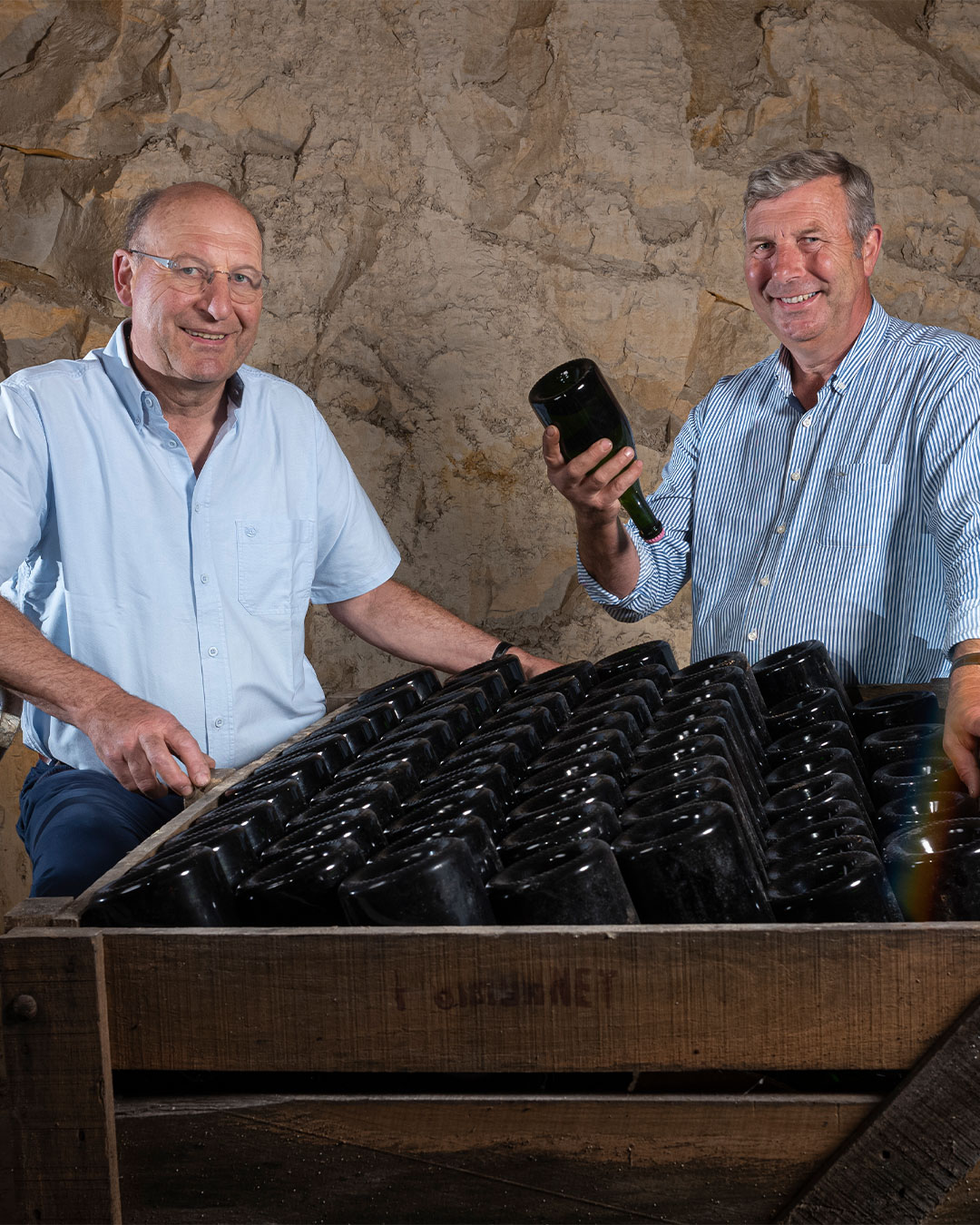 Zwei Männer stehen in einem Raum mit einer steinernen Wand und einer Holzplatte voller dunkelgrüner Flaschen. Einer von ihnen hält eine Flasche in der Hand und lächelt.