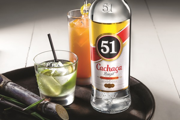 Eine Flasche Cachaça 51 steht auf einem Tablett. Daneben befindet sich ein Glas mit Eis und Limette, sowie ein weiteres Glas mit orangenem Getränk. Zuckerrohr liegt ebenfalls auf dem Tablett.