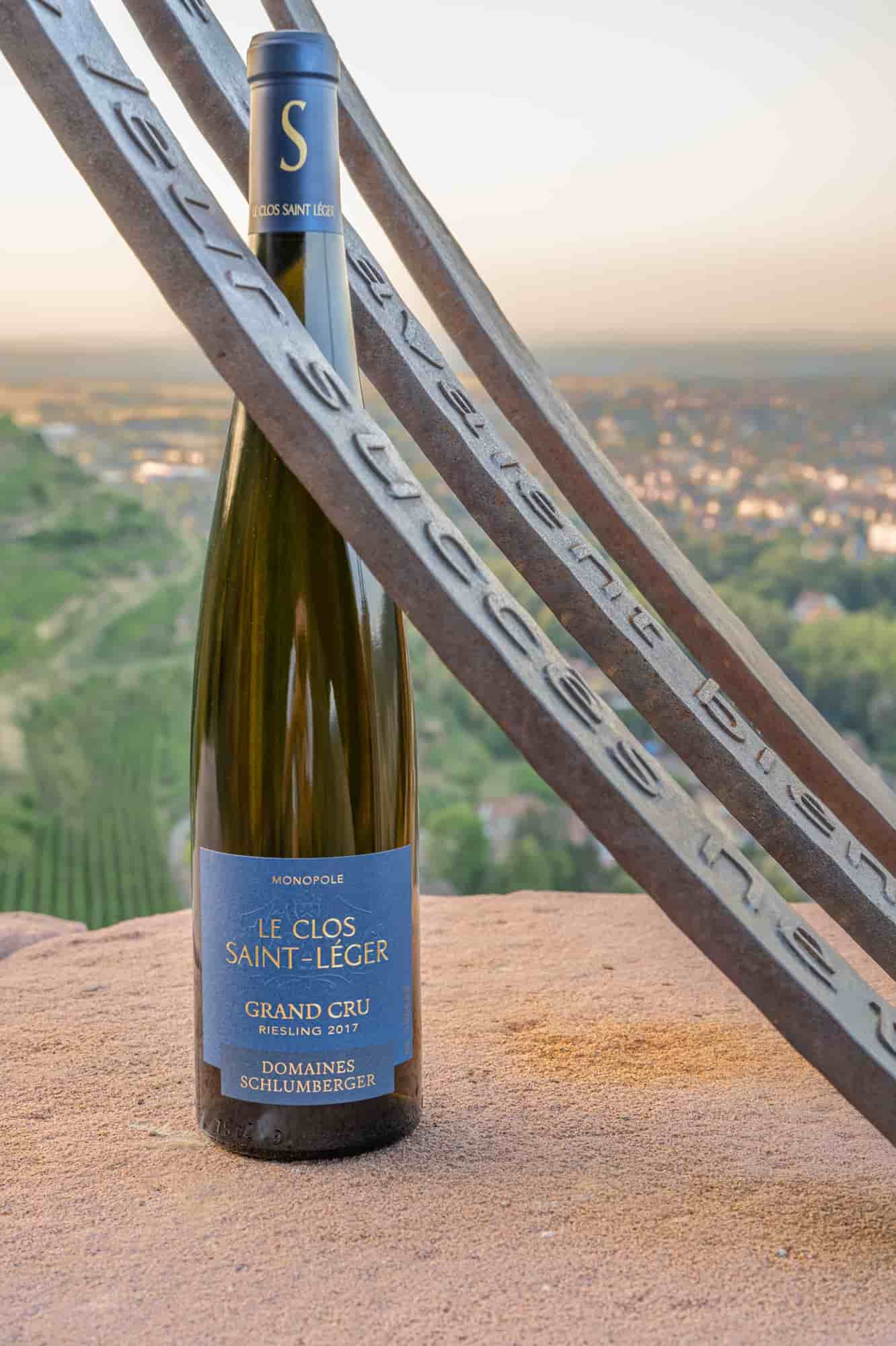 Eine Weinflasche steht auf einem steinigen Untergrund, umgeben von Metallteilen. Im Hintergrund erstrecken sich grüne Weinberge und eine ländliche Landschaft bei Sonnenuntergang.