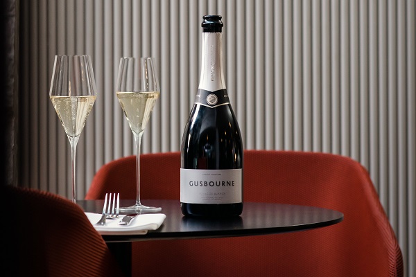 Gusbourne_Blanc_de_Blancs_600 Eine Flasche Gusbourne-Sekt steht auf einem kleinen Tisch. Neben der Flasche befinden sich zwei Gläser mit Sekt, auf einem weißen Servietten liegt ein Besteck. Der Hintergrund zeigt eine gestreifte Wand und rote Stühle.