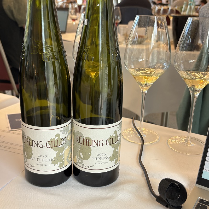 Zwei Weinflaschen stehen nebeneinander auf einem Tisch, beschriftet mit -Kühling-Gillot-, Jahrgang 2025. Daneben stehen zwei Weingläser mit hellgelbem Wein. Im Hintergrund sind weitere Gläser und eine Tischdecke sichtbar.