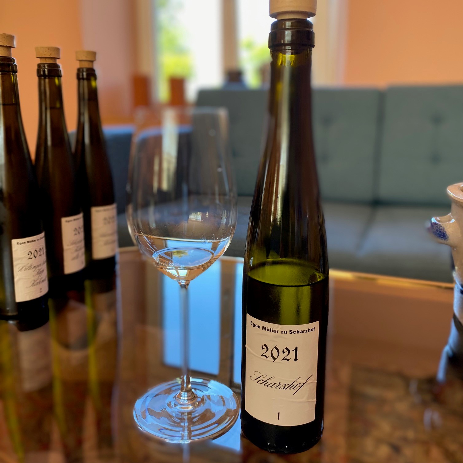 Eine Flasche Wein mit dem Etikett -2021 Scharzhof- steht neben einem leeren Weinglas auf einem Tisch. Im Hintergrund sind weitere Weinflaschen sichtbar und ein grünes Sofa.