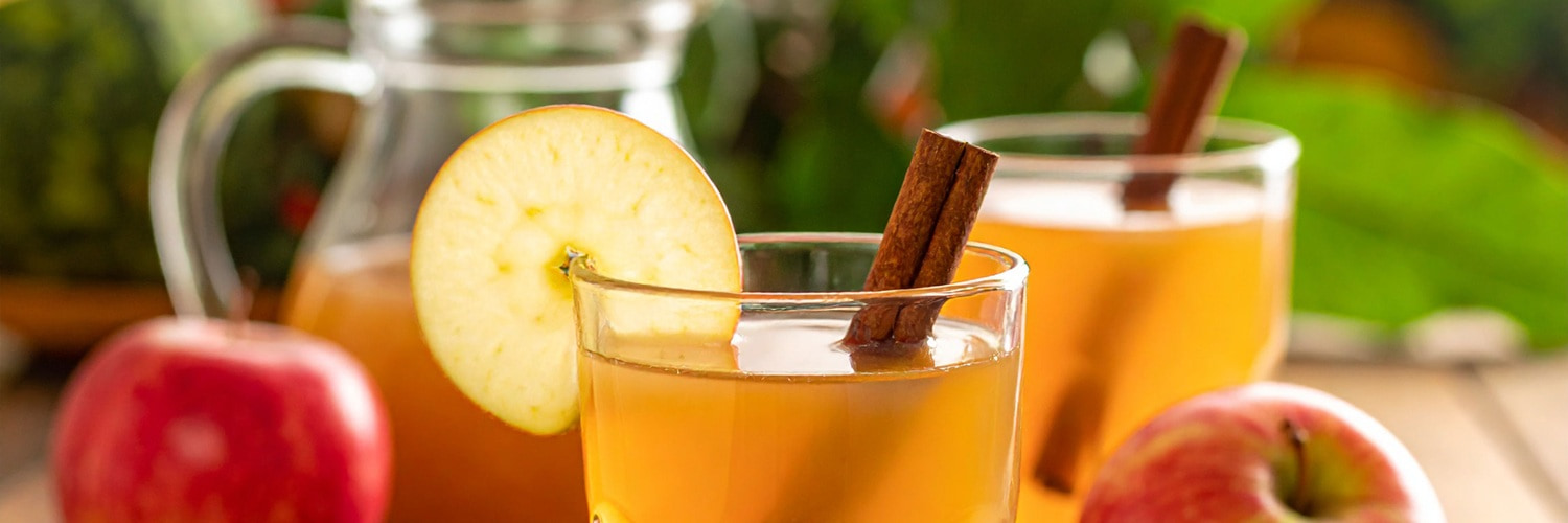 Eine weiße Tasse mit einem schwarzen Rand steht auf einem gestrickten Stoff. Dampfwölkchen steigen auf, während rote Beeren und trockene Blätter daneben liegen.