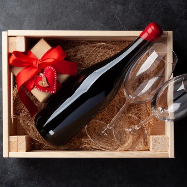 Ein Holzgeschenkbox enthält eine Flasche Rotwein, zwei Weingläser und ein Päckchen mit rotem Geschenkband, umgeben von Holzspänen. Der Kontext ist schlicht und elegant gestaltet.