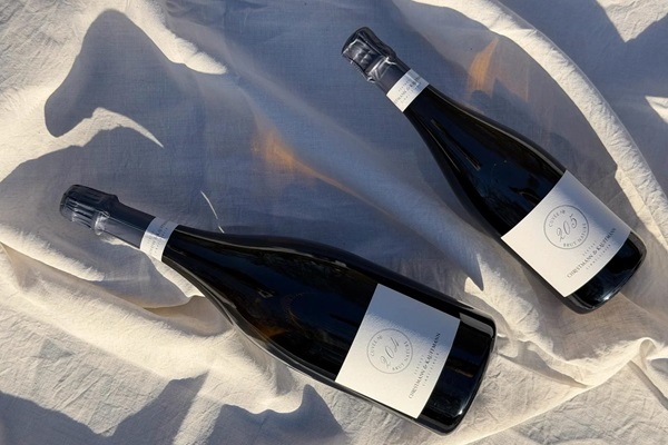 Zwei Flaschen Sekt liegen auf einem creméfarbigen Tuch. Sie sind dunkel und elegant gestaltet, mit weißen Etiketten, die Details über den Sekt zeigen.