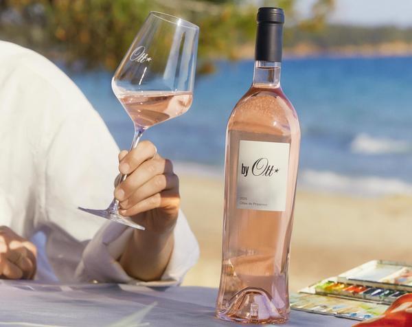 Ein Glas Roséwein wird von einer Person gehalten, während eine Flasche -by Ott*- auf einem Tisch steht. Der Hintergrund zeigt eine Küstenlandschaft mit Wasser und Bäumen.
