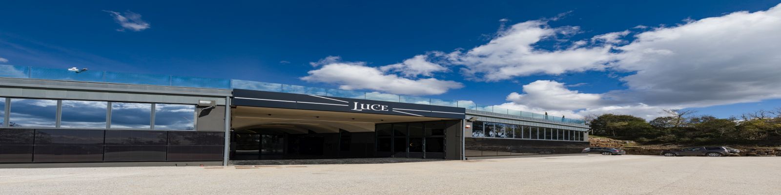 Tenuta Luce Ein modernes Gebäude mit einer großen Glasfront steht vor einem klaren blauen Himmel. Der Eingangsbereich ist markant mit dem Schriftzug -Luce- gestaltet, umgeben von Parkplatz und Bäumen.