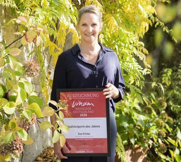 Eine Frau mit blonden Haaren steht lächelnd vor buntem Laub. Sie hält eine Auszeichnung in der Hand, die -Aufsteigerin des Jahres- für Eva Fricke zeigt.