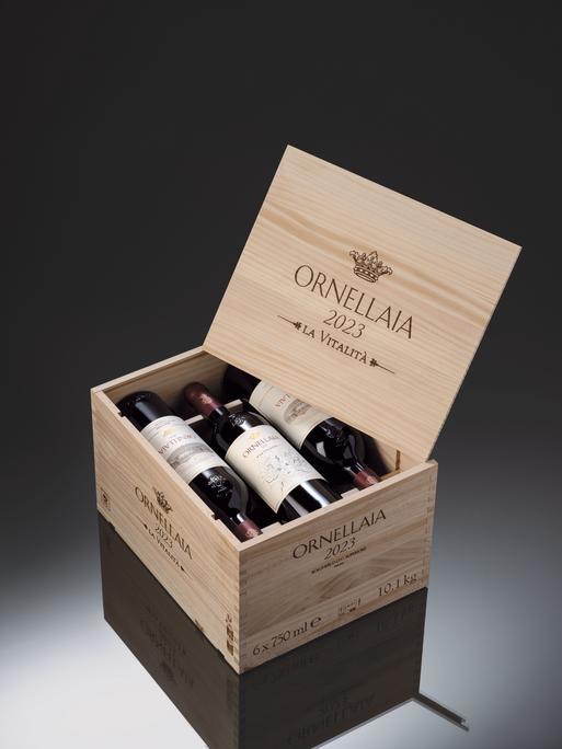Eine offenstehende Holzkiste enthält sechs Flaschen Wein, mit Etiketten. Die Kiste trägt die Aufschrift -ORNELLAIA 2023 – LA VITALITÀ- und steht auf einer reflektierenden Oberfläche.