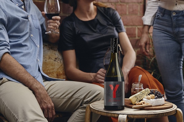 Shiraz-Y-600 Eine Weinflasche mit einem auffälligen Etikett steht auf einem Tisch. Menschen genießen Wein und Snacks, während sie in einer entspannten Umgebung mit Ziegelwand sitzen.