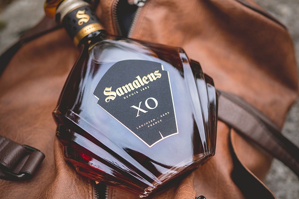 Ein eleganter, flaschenförmiger Cognac mit dem Etikett -Samalens- XO- liegt auf einer braunen Ledertasche. Die Flasche hat eine facettierte Form und glänzt im Licht.