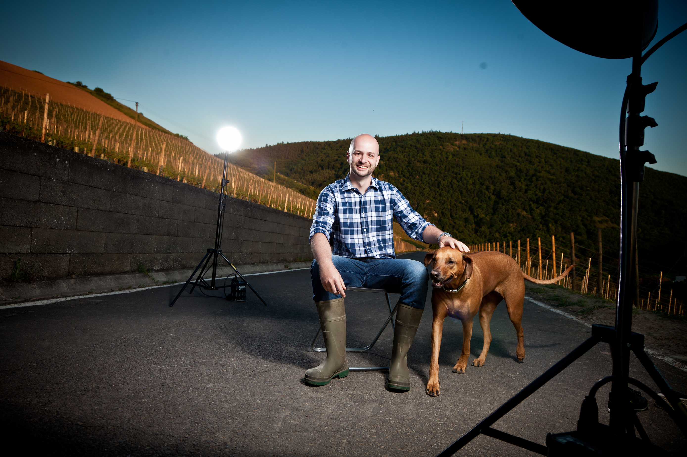 Ein Mann sitzt auf einem Stuhl und lächelt, während ein Hund neben ihm steht. Im Hintergrund sind Weinreben und eine beleuchtete Fotoständer zu sehen, unter einem klaren, blauen Himmel.