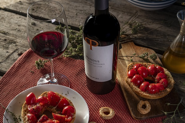 Ein Glas Rotwein steht neben einer Flasche mit dem Etikett -Mandus-. Auf einem Teller sind Brote mit Tomaten und Kräutern angerichtet, während ein rustikaler Tisch die Szenerie umrahmt.