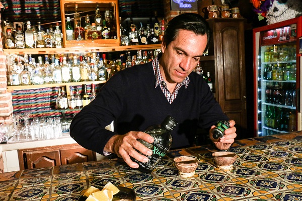 Ein Mann schüttet in einer Bar Tequila in zwei traditionelle Schnapsgläser. Hintergrund ist eine gut gefüllte Bar mit verschiedenen Spirituosen. Auf dem Tisch liegen Limettenstücke.