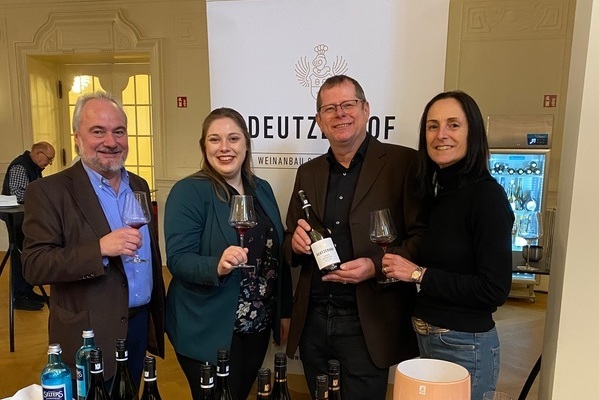 Eine Gruppe von vier Personen steht zusammen und hält Weingläser in der Hand. Im Hintergrund ist ein Banner mit dem Namen -DEUTZ- zu sehen, und mehrere Flaschen Wein stehen auf einem Tisch.