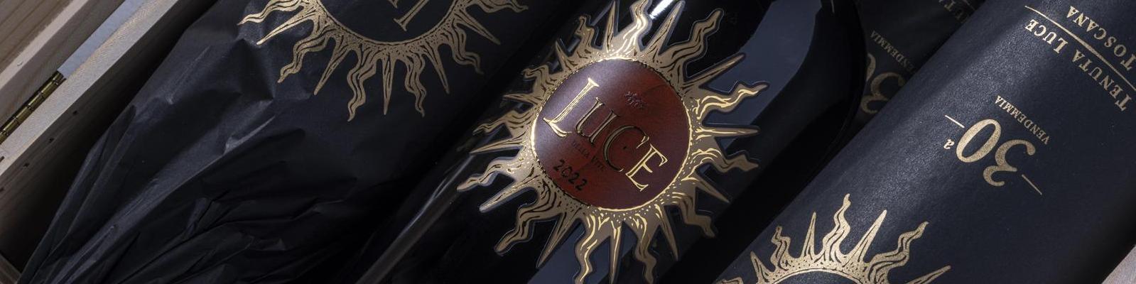 Drei Flaschen Wein sind in schwarzem Papier mit goldenen Sonnendesign verpackt. Auf den Etiketten steht -Luce- und -2022-, umgeben von goldenen Strahlen.