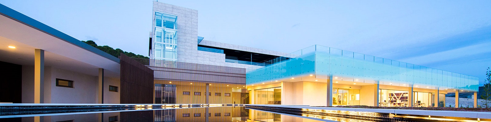 Aalto Ein modernes Gebäude mit großen Fenstern und einer gläsernen Fassade steht an einem ruhigen Ort. Es reflektiert sich im Wasserbecken davor und wird von einer leicht bewölkten Abenddämmerung umgeben.