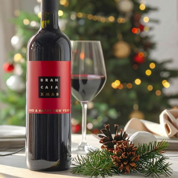 Eine Flasche Rotwein steht auf einem Tisch, daneben ein Glas Wein. Im Hintergrund ist ein Weihnachtsbaum mit Lichtern und Dekorationen zu sehen, umgeben von Tannenzweigen und einer Zapfen.