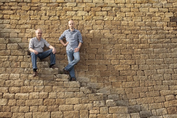 Zwei Männer sitzen und stehen auf einer steinernen Treppe, die an einer großen, strukturierten Steinmauer entlangführt. Die Mauer hat eine gelbliche Farbe und die Umgebung ist hell.