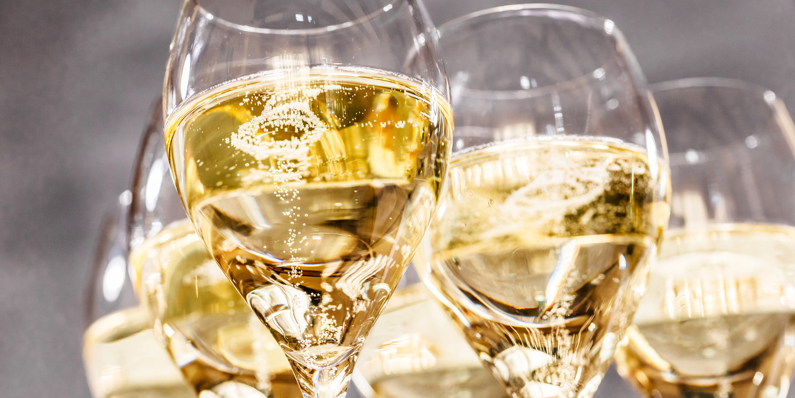 Sechs Gläser mit schaumigem Weißwein stehen eng beieinander. Blasen steigen im Getränk auf, während der unscharfe Hintergrund eine elegante Atmosphäre erzeugt.