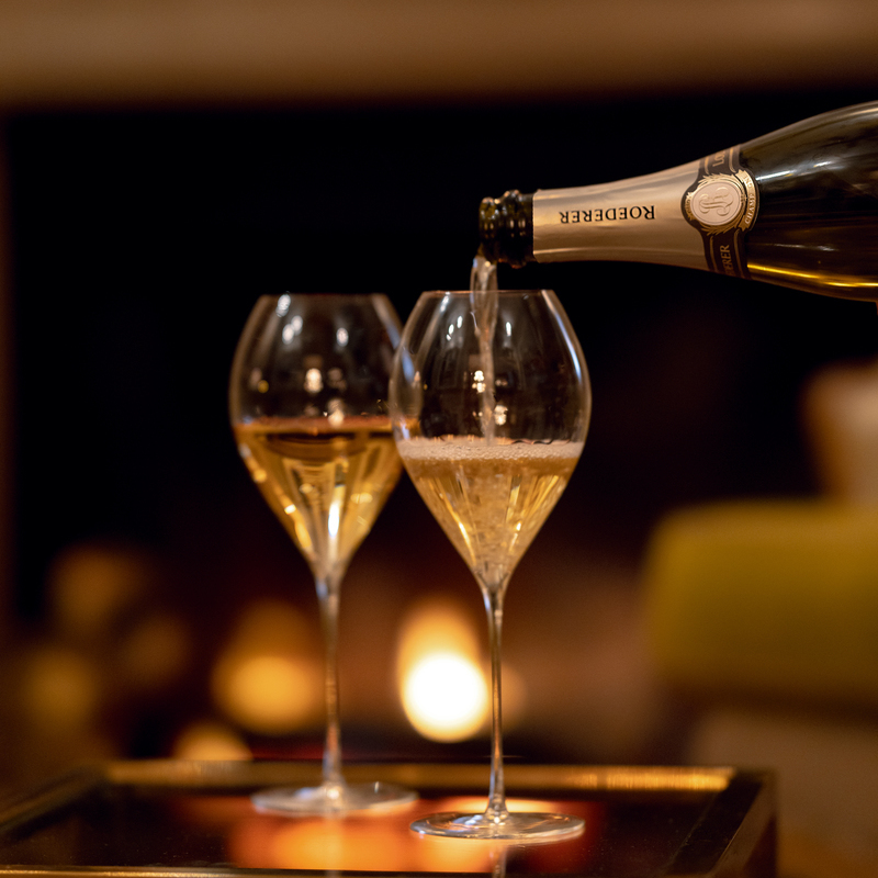 Ein Sektglas wird gefüllt, während ein zweites Glas daneben steht. Im Hintergrund flackert ein Kaminlicht, das eine gemütliche Atmosphäre schafft.