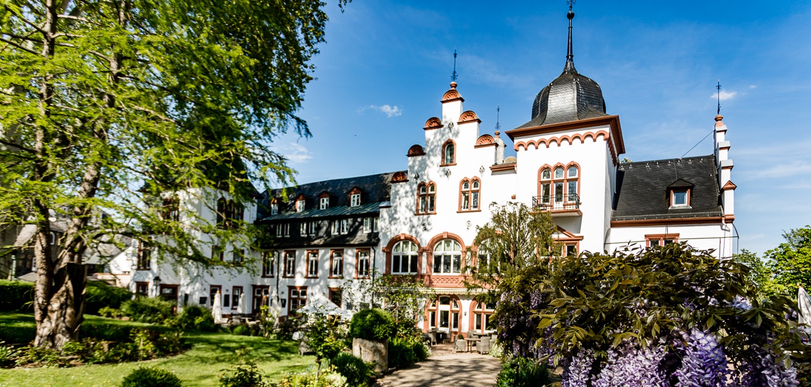Ein historisches Gebäude mit rotem und weißem Putz, geschmückt mit Türmchen und Erkern. Umgeben von einer grünen Landschaft und blühenden Pflanzen, strahlt es einen einladenden Charme aus.
