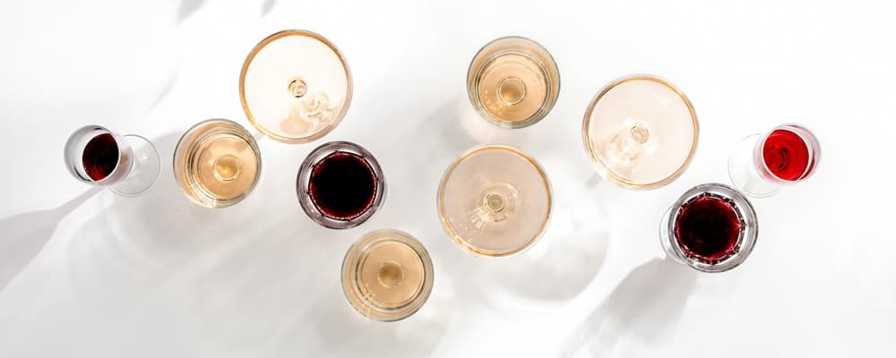 Sechs Weingläser mit verschiedenen Weinsorten (rot, rosé, weiß) liegen chaotisch auf einer grauen Oberfläche. Ein Korkenzieher und ein Korken sind daneben platziert.