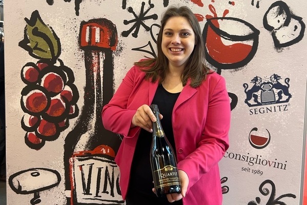 Eine Frau in einem pinken Blazer hält eine Flasche Sekt. Im Hintergrund sind Weintrauben, Gläser und die Schriftzüge -VIN- und -consiglionvini seit 1989- zu sehen.