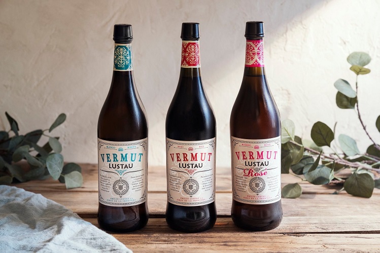 Drei Flaschen Vermut Lustau stehen nebeneinander auf einem Holztisch. Jede Flasche hat ein einzigartiges Etikett und eine unterschiedliche Farbe. Im Hintergrund sind einige grüne Blätter und sanftes Licht sichtbar.