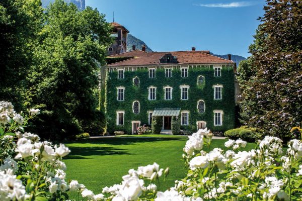 Ein mehrstöckiges Gebäude, mit grüner Pflanzenverkleidung, steht in einem gepflegten Garten. Vorne blühen weiße Rosen, während hohe Bäume und Berge die Szenerie im Hintergrund ergänzen.