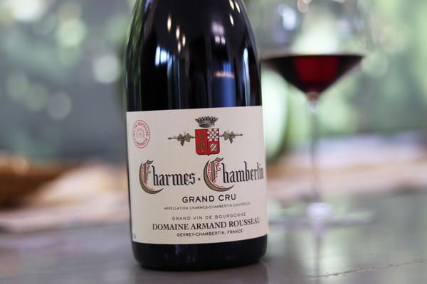 Eine Flasche Charmes-Chambertin Grand Cru Wein steht auf einem Tisch. Im Hintergrund ist ein Weinglas mit rotem Wein und eine verschwommene, grüne Umgebung zu sehen.