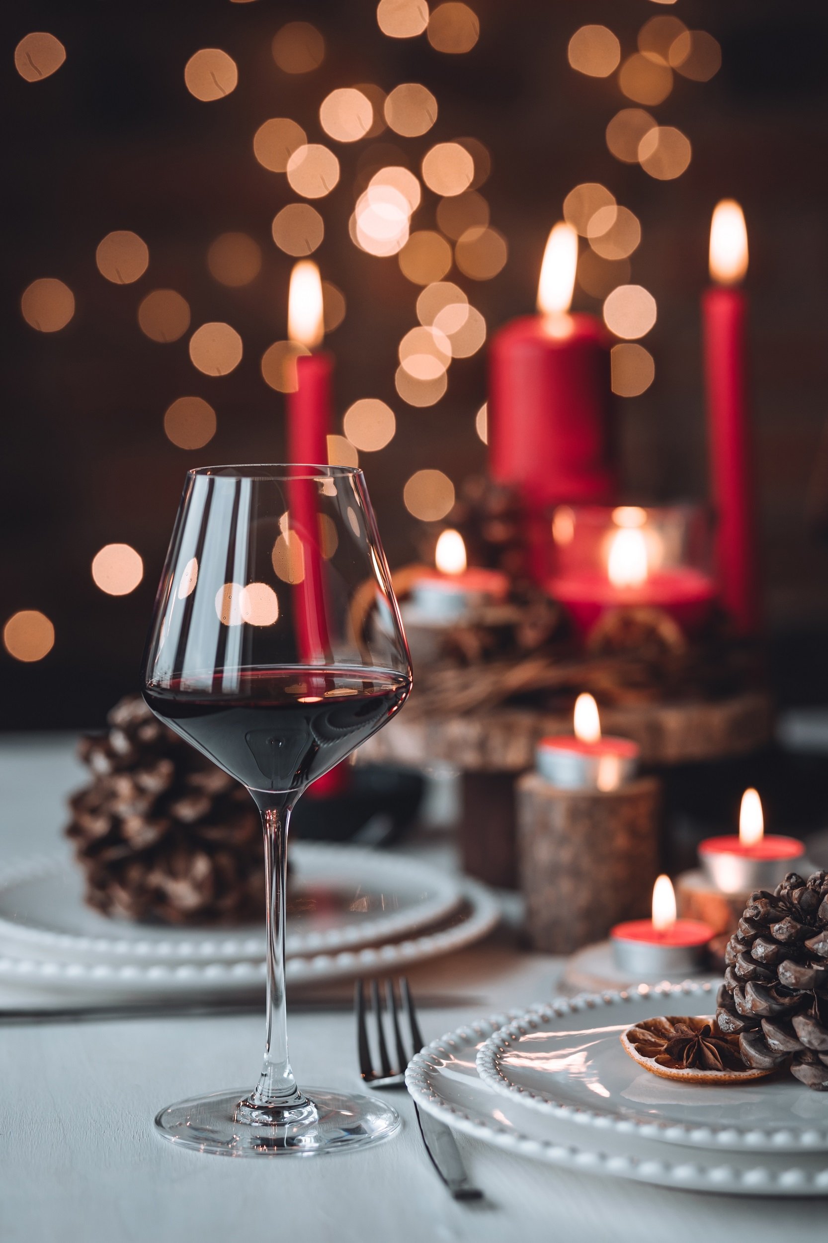 Ein Weinglas mit rotwein steht auf einem festlich gedeckten Tisch. Im Hintergrund leuchten rote Kerzen und weihnachtliche Lichter, umgeben von Tannenzapfen und dekorativen Platten.