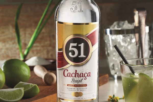 Eine Flasche Cachaca 51 steht im Vordergrund. Daneben liegen Limetten und ein Mörser. Im Hintergrund sind Eiswürfel und ein Glas mit einem Cocktail sichtbar, während grüne Blätter dekorativ angeordnet sind.