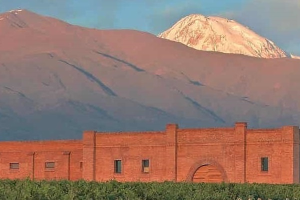 Eine rote Mauerstruktur steht im Vordergrund, umgeben von Weinreben. Im Hintergrund erhebt sich ein schneebedeckter Berg unter einem klaren Himmel.
