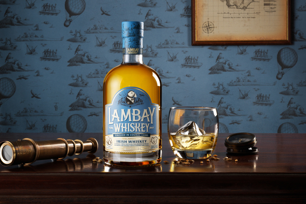 Eine Flasche Lambay Whiskey steht auf einem Holztablett, daneben ein Glas mit Whiskey und Eiswürfeln. Im Hintergrund sind alte Seekarten und Luftballons auf blauer Tapete zu sehen.