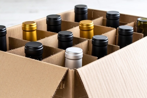 Eine Kartonschachtel enthält mehrere Flaschen mit unterschiedlichen Kappenfarben, darunter Schwarz, Silber und Gold. Die Flaschen sind ordentlich in Fächern angeordnet, auf einem hellen Untergrund.