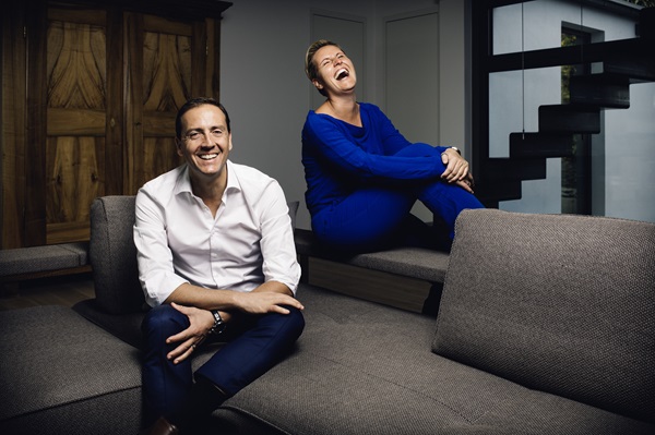 Ein Mann sitzt auf einem grauen Sofa und lächelt, während eine Frau in blauem Outfit fröhlich hinter ihm sitzt und lacht. Der Hintergrund zeigt Fenster und eine Holzschrankwand.