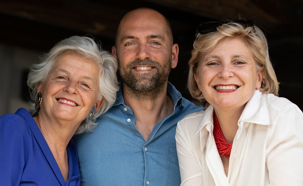 Drei Personen lächeln fröhlich in die Kamera. Sie stehen eng zusammen, umgeben von einem rustikalen Hintergrund, der eine freundliche, informelle Atmosphäre schafft.