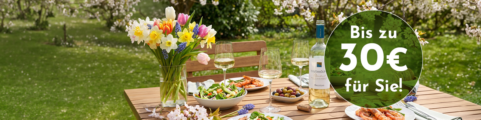 Ein festlich gedeckter Tisch im Freien mit Blumen, Weingläsern und verschiedenen Gerichten. Im Hintergrund blühende Bäume und grüne Wiese. Text: -Bis zu 30€ für Sie!-