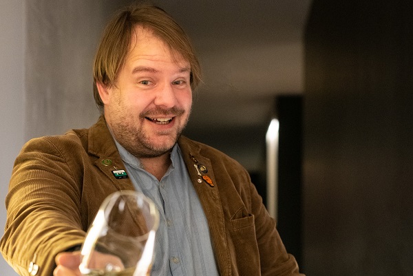 Ein lächelnder Mann mit kurzen, hellen Haaren trägt einen braunen Cord-Blazer und hält ein Glas in der Hand. Er steht in einem modernen und schlichten Innenraum.