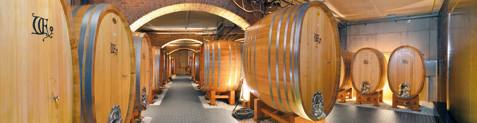 Große Holzfässer stehen nebeneinander in einem Keller mit Ziegelwänden. Der Boden ist mit Fliesen belegt, und die Fässer sind durch sanfte Beleuchtung hervorgehoben.