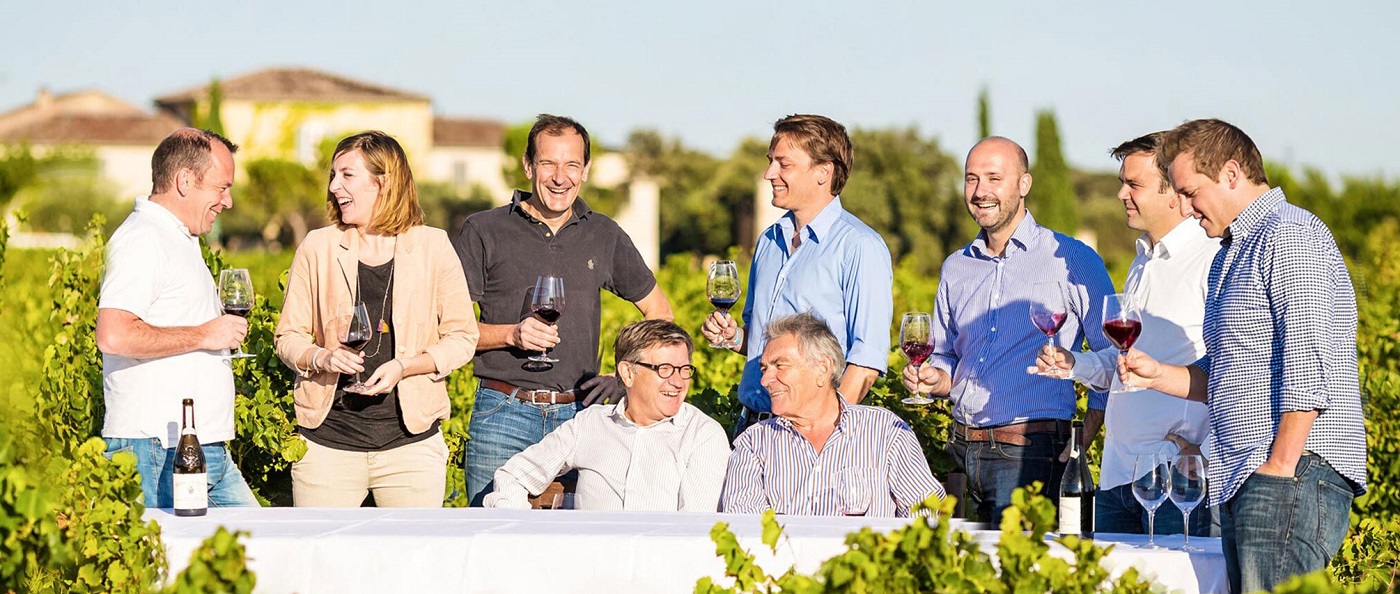 Eine Gruppe von acht Personen steht in einem Weinberg, lacht und hält Weingläser. Einige sitzen an einem Tisch, eine Flasche Wein steht auf dem Tisch. Im Hintergrund sind Weinreben und ein Gebäude zu sehen.