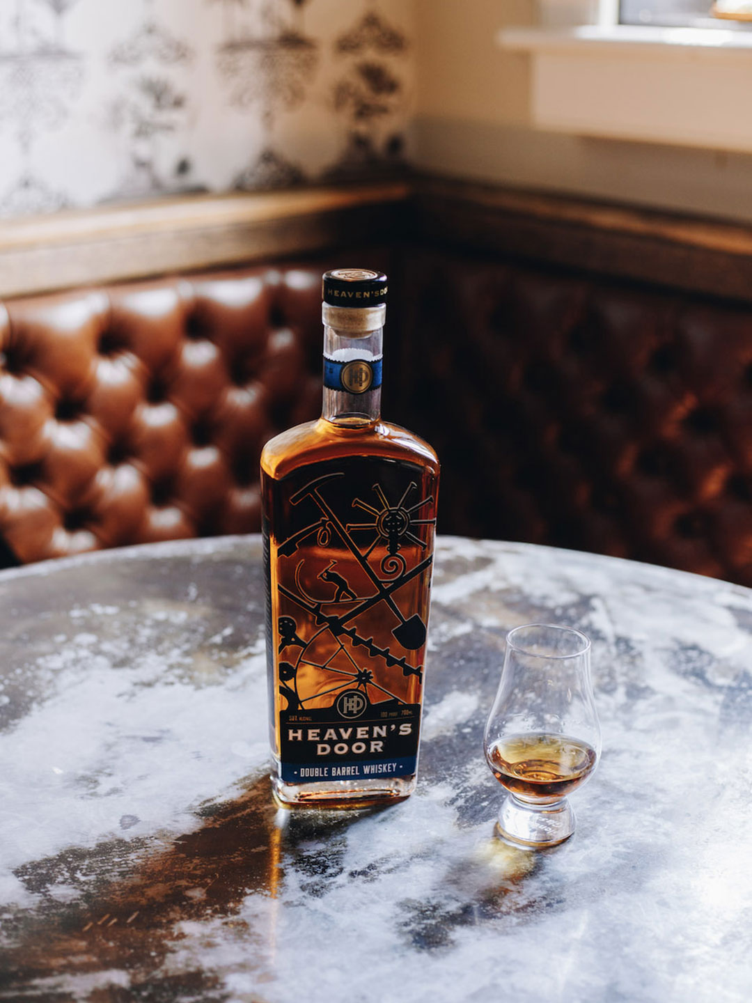 Eine Flasche -Heaven-s Door- Whiskey steht auf einem glänzenden, abgenutzten Tisch. Neben der Flasche befindet sich ein Glas mit Whiskey, im Hintergrund sind gepolsterte Wände sichtbar.