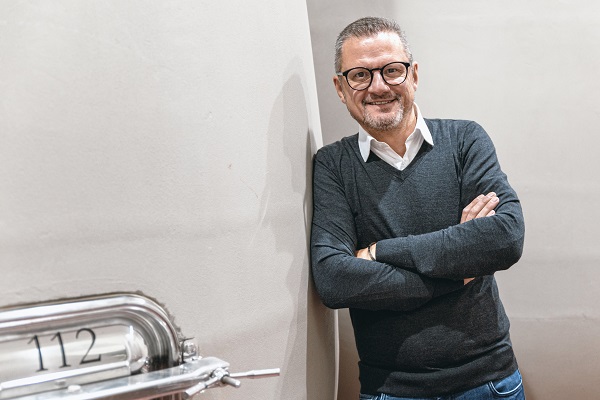 Ein Mann mit Brille und einem lächelnden Gesicht lehnt an einer Wand. Er trägt einen dunkelgrauen Pullover über einem weißen Hemd. Neben ihm steht ein metallisches Objekt mit der Nummer 112.