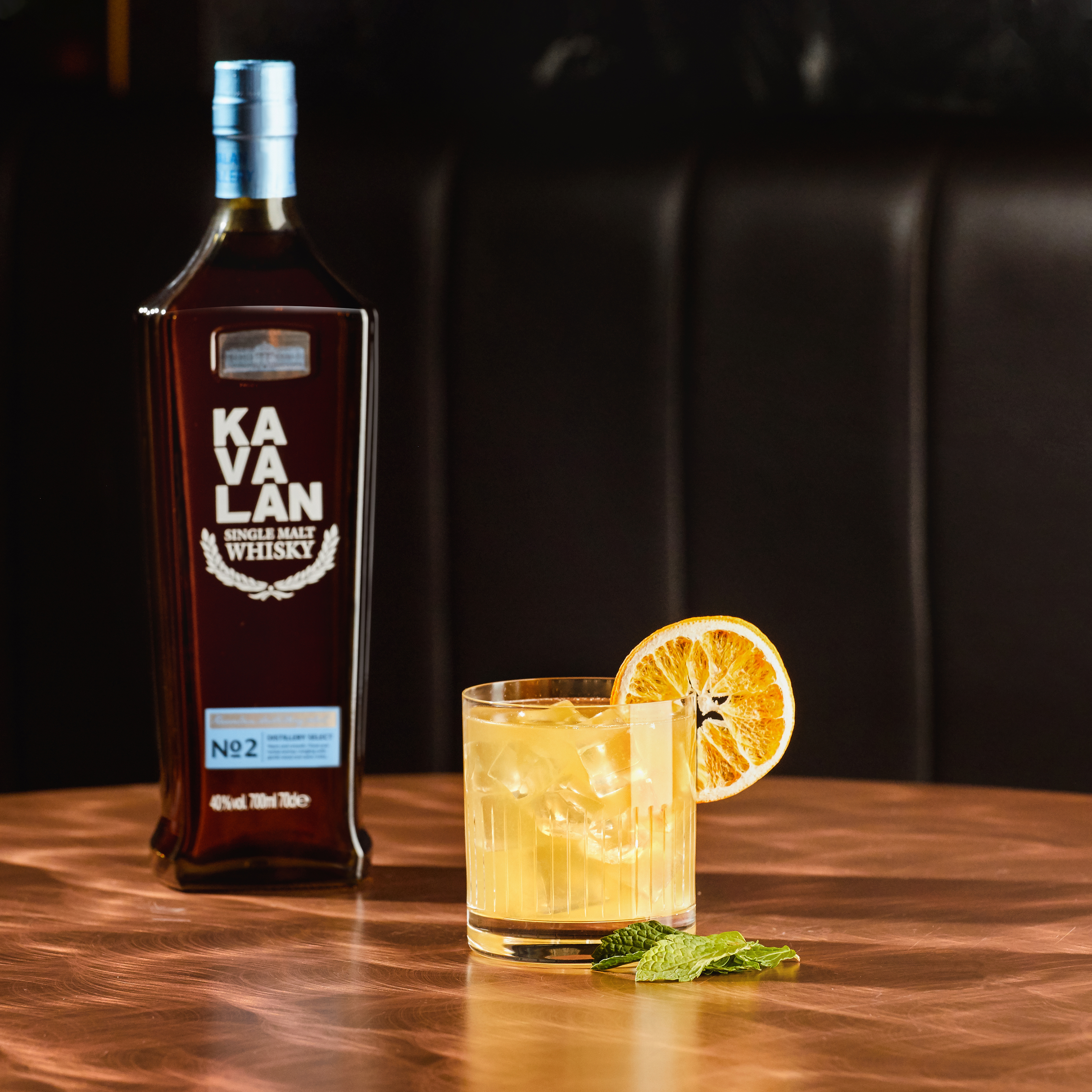 Eine Flasche Kavalan Single Malt Whisky steht neben einem Glas mit Eiswürfeln und einem dekorativen Orangenscheibchen. Der Tisch hat eine glänzende Kupferoberfläche.