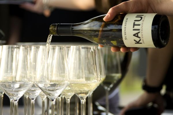 Ein Glas gefüllt mit Weißwein wird aus einer Flasche mit der Aufschrift -Kaitui- nachgeschenkt. Im Hintergrund sind weitere Gläser sichtbar, während eine Hand die Flasche hält.