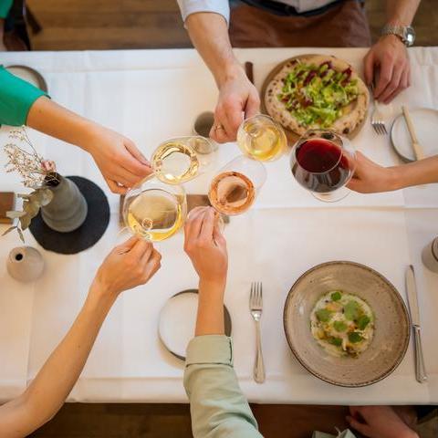 Mehrere Hände heben Gläser mit Getränken in einem Restaurant. Auf dem Tisch liegen verschiedene Speisen und Besteck neben einigen dekorativen Elementen. Es herrscht eine gesellige Atmosphäre.