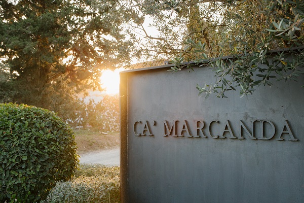 Ein elegantes Schild mit der Aufschrift -CA’ MARCANDA- steht am Wegesrand, umgeben von grünen Sträuchern. Im Hintergrund strahlt die Sonne, die den Morgenhimmel erleuchtet.